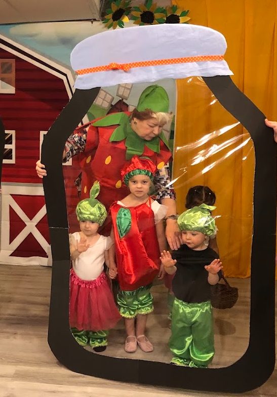 Children in strawberry and cabbage costumes posed with a teacher in a giant mason jar photo prop